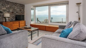 A modern living room with a stone wall, a wooden entertainment unit, a large window with a view of the sea, and a grey sofa with blue cushions. - Awel Y Mor (Criccieth)
