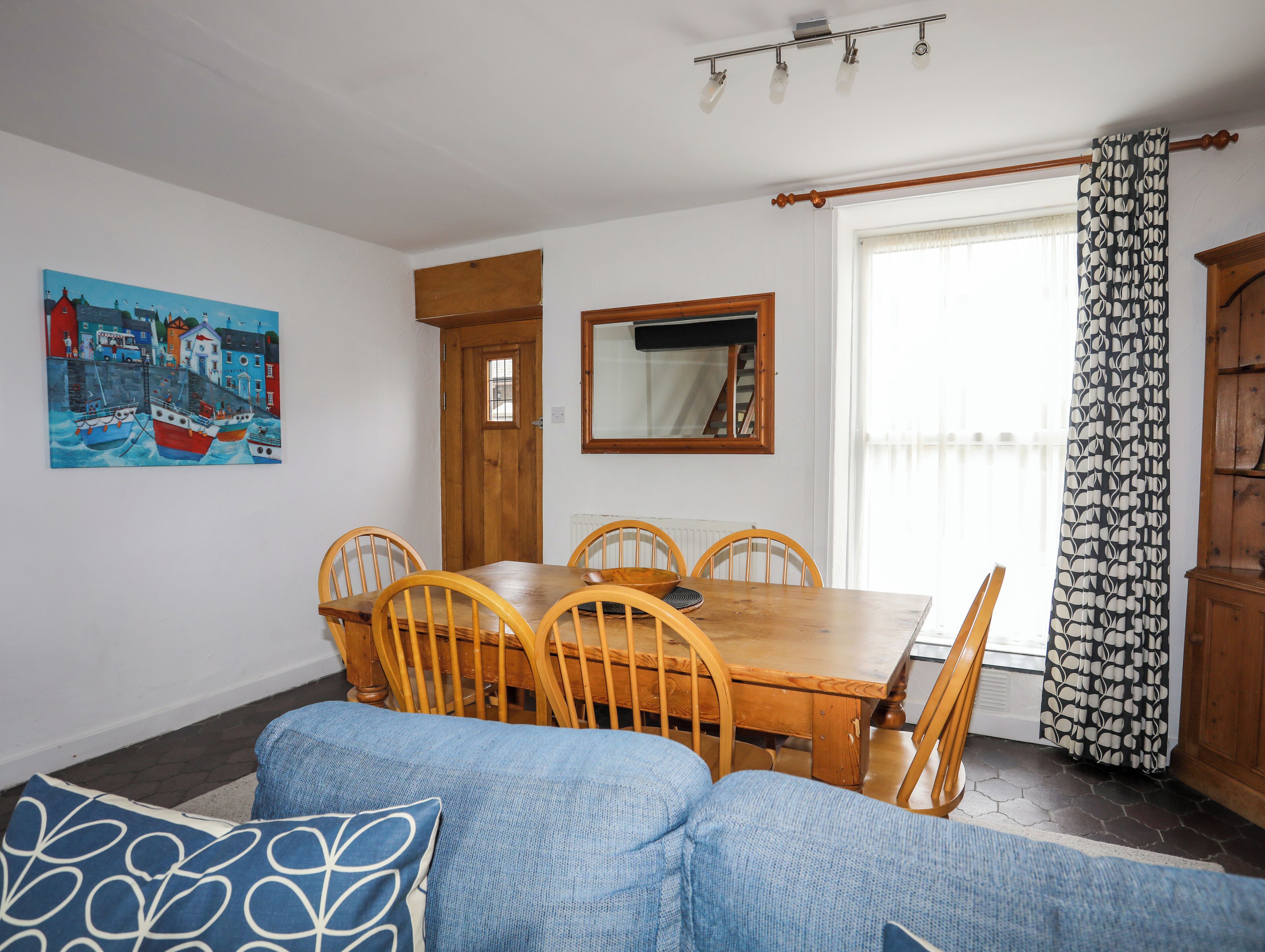 A dining area with a wooden table and chairs, a blue sofa, a painting of boats, a window with a curtain, and a mirror on the wall.