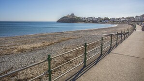 A coastal promenade with a pebble beach, a metal railing, and a distant hill with a structure on top. - Awel Y Mor (Criccieth)
