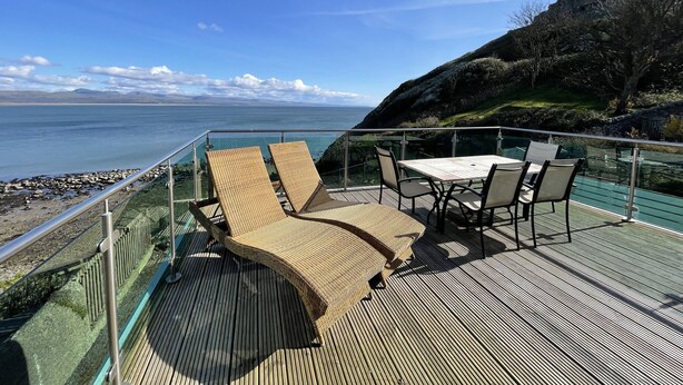 A deck with wicker chairs and a table overlooking a rocky shoreline and the sea. - Awel Y Mor (Criccieth)
