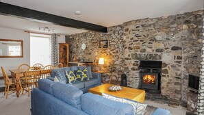 A cozy living room with a stone wall, a fireplace, a blue sofa, and a dining area with wooden furniture. - Awel Y Mor (Criccieth)