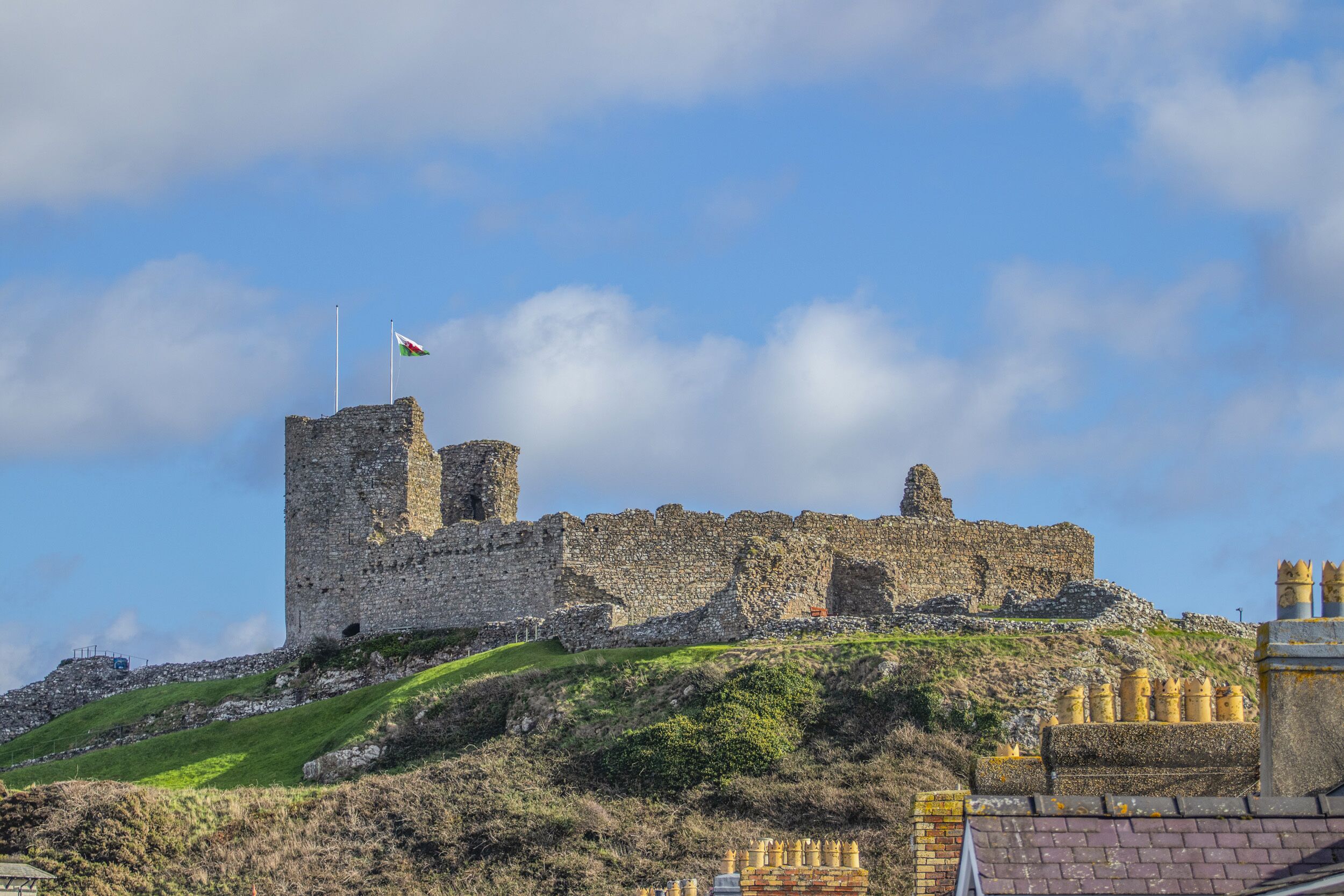Um castelo de pedra histórico com uma bandeira no topo, situado numa colina gramada sob um céu limpo.