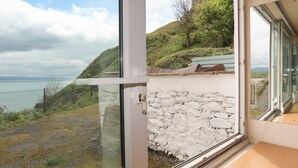 A room with a large window offering a view of a stone wall, a grassy hill, and a distant body of water. - Awel Y Mor (Criccieth)