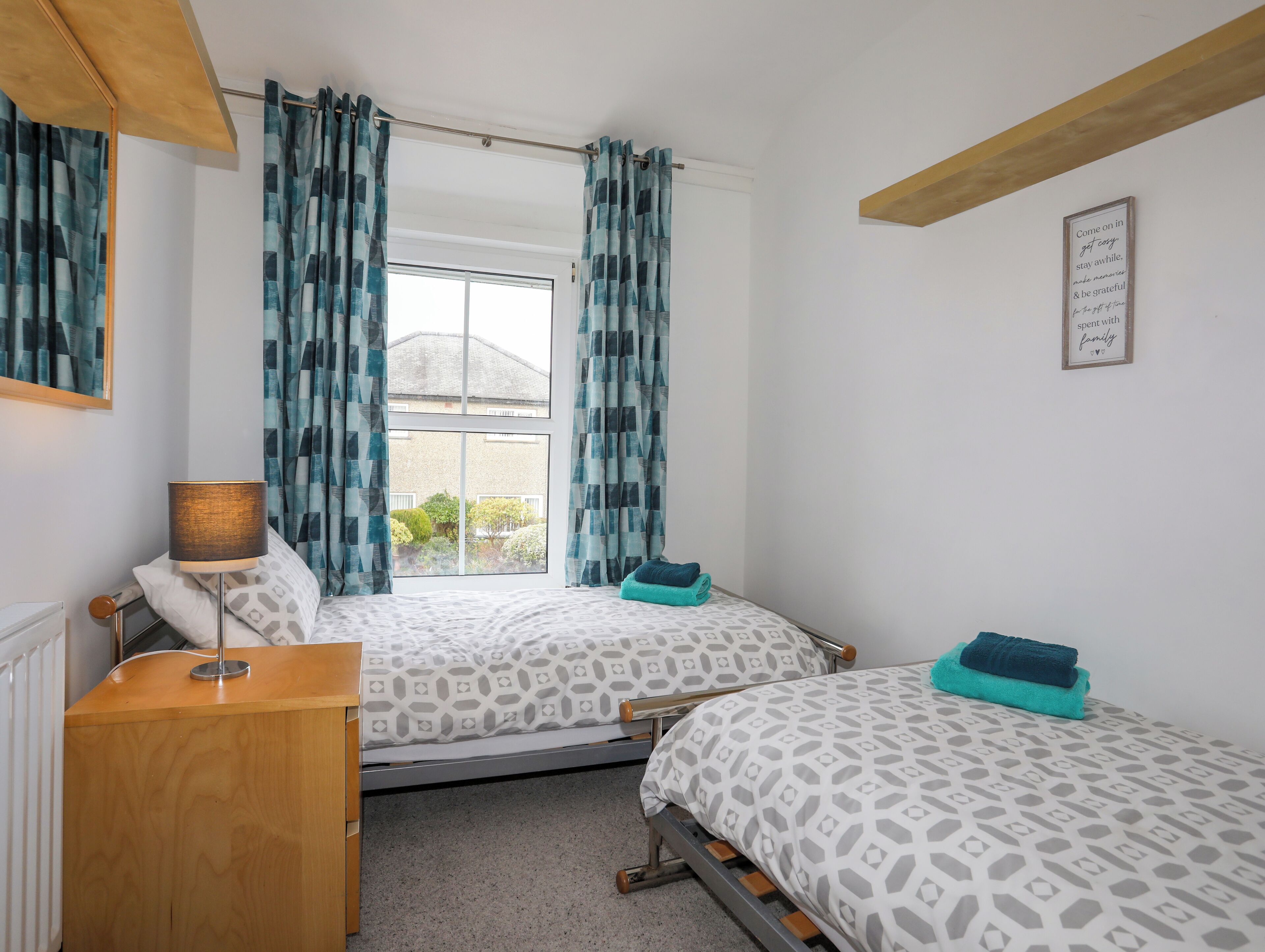 A bedroom with two beds, a window with curtains, a bedside table with a lamp, and a framed quote on the wall.