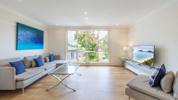 Uma sala de estar moderna com um sofá, uma mesa de centro de vidro e uma televisão exibindo uma cena de praia.