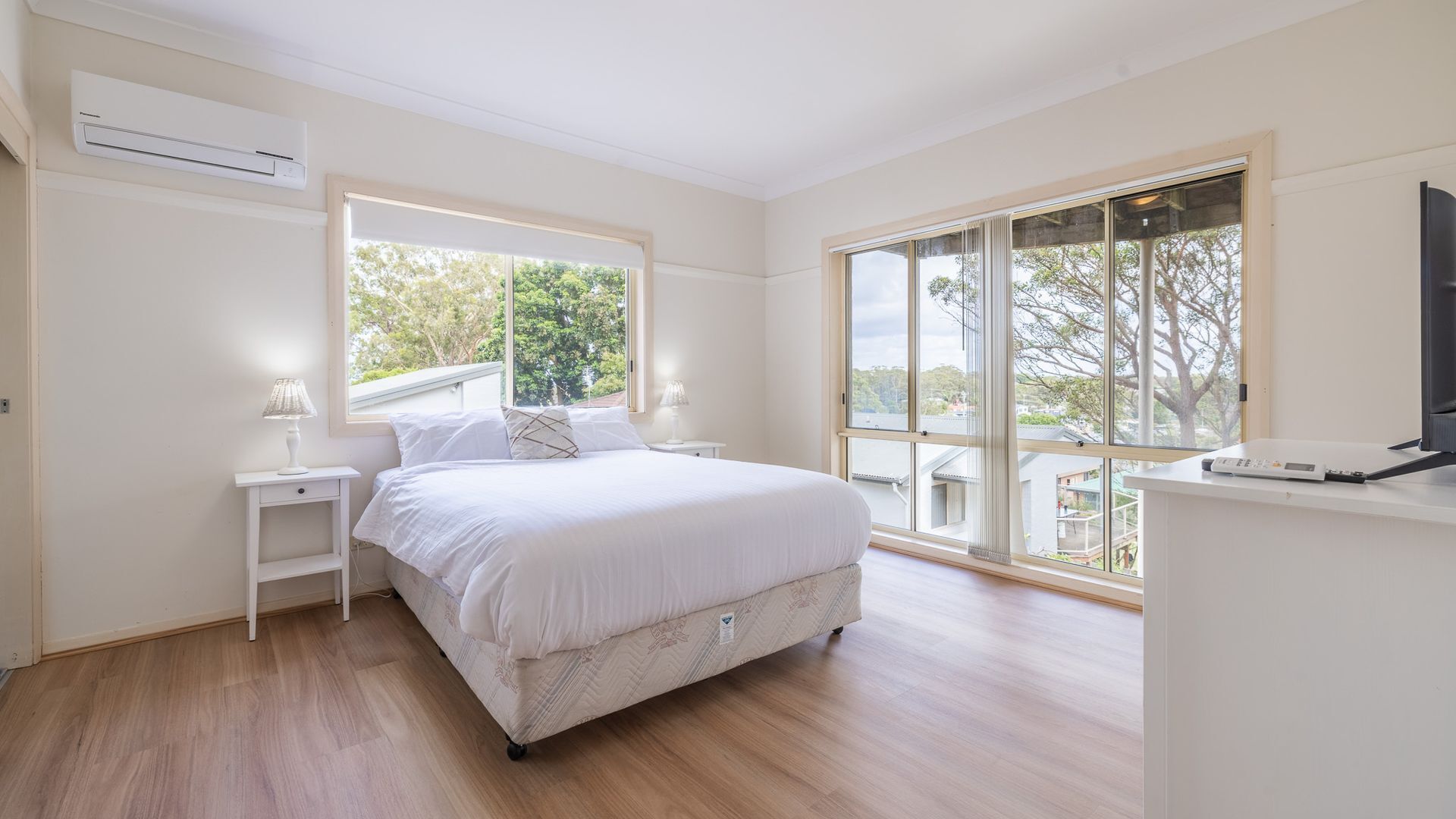 Une chambre à coucher comprenant un lit, des tables de chevet, un appareil de climatisation, une télévision et de grandes fenêtres donnant sur des arbres et des immeubles.
