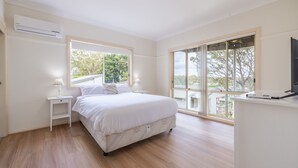Quarto com cama, mesas de cabeceira, ar condicionado, televisão e grandes janelas com vista para árvores e edifícios.