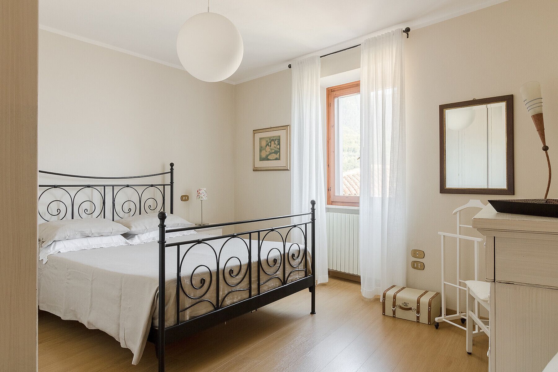 Een slaapkamer met een bed, een dressoir, een spiegel, een raam met gordijnen en een hanglamp.