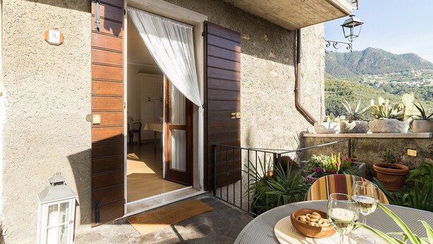 A balcony with a view of mountains, a wooden door, a small table with a bowl of snacks and glasses, and potted plants. - Corte Trepée, in the old town of Piovere (Tignale)