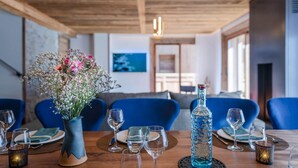 Un coin repas avec une table en bois dressée pour quatre personnes, des chaises bleues, un vase avec des fleurs et une bouteille d’eau.