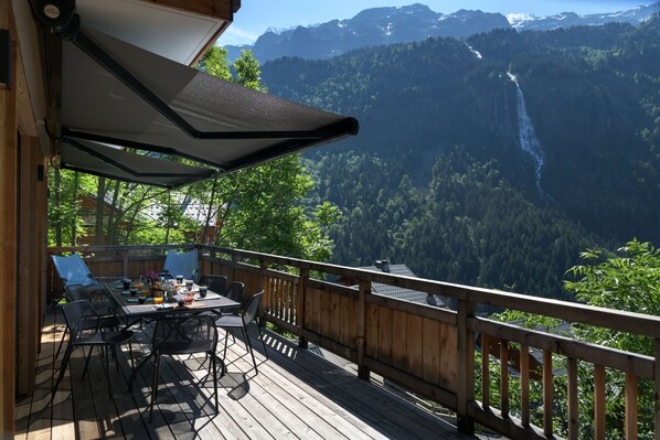 A wooden deck with a table and chairs, overlooking a mountainous landscape with a waterfall.
