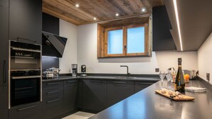 A modern kitchen with dark cabinetry, a built-in oven, a coffee maker, and a wooden window above the sink. - "Refuge des Sens", Chalet "Quiétude" (Vaujany)