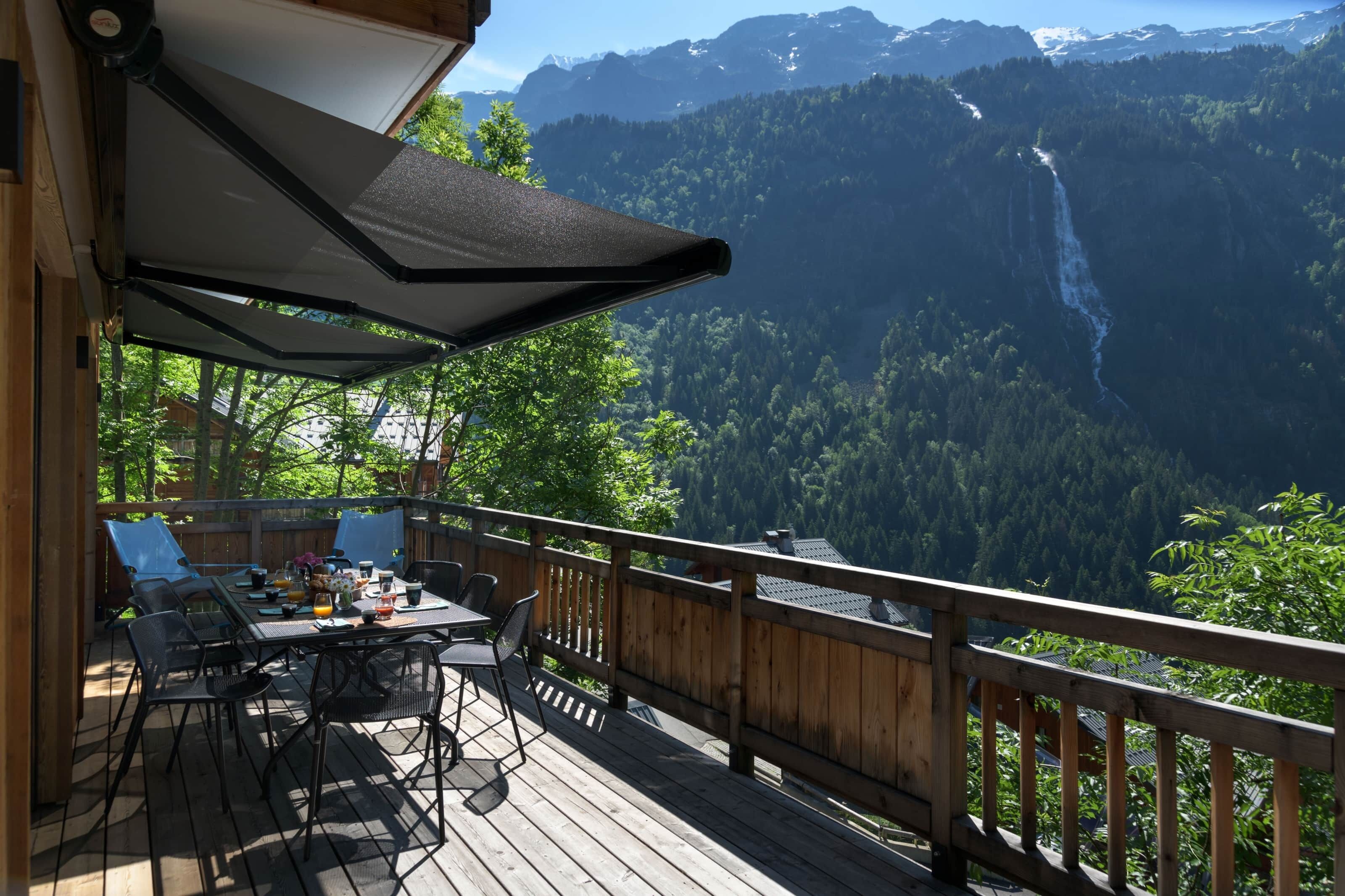 A wooden deck with a table and chairs, overlooking a mountainous landscape with a waterfall.