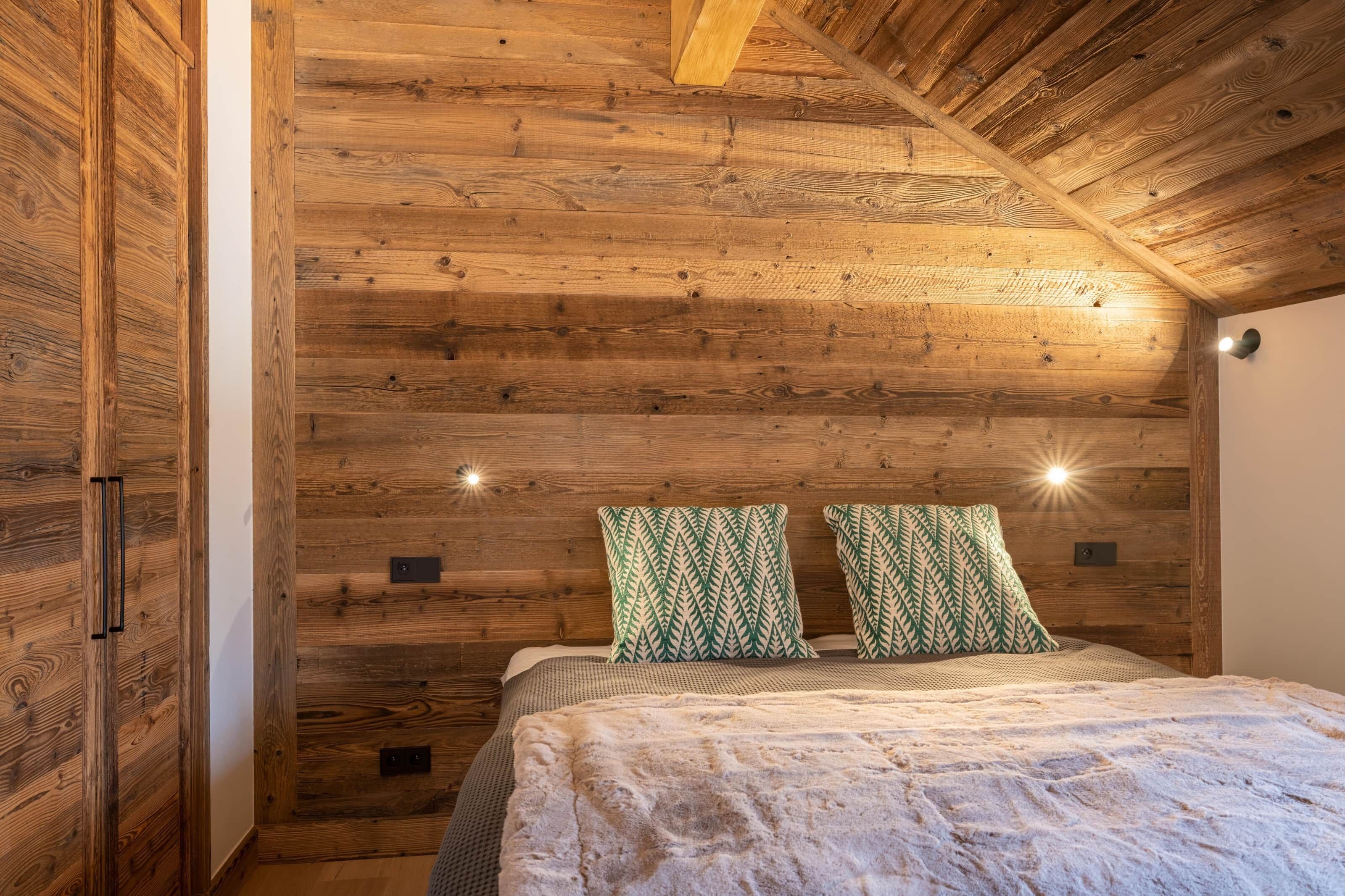 Un dormitorio con paredes y techo de madera, una cama con almohadas estampadas y dos luces fijadas a la pared.