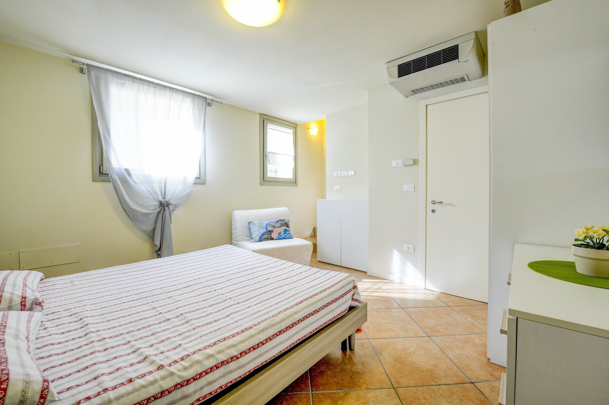 Un dormitorio con cama, ventana con cortinas, mesita de noche con lámpara, armario y unidad de aire acondicionado.