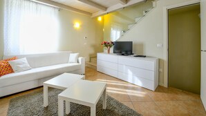 Una sala de estar con un sofá blanco, una mesa de centro, un televisor sobre un mueble blanco y una escalera que lleva a una puerta.