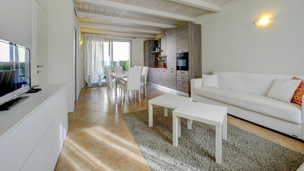 Una sala de estar moderna con un sofá blanco, una mesa de centro y un televisor sobre un mueble bajo. Hay una zona de cocina con armarios y una mesa de comedor con sillas.