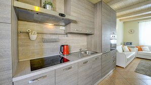 Una cocina moderna con estufa integrada, lavamanos y campana extractora de acero inoxidable. Hay una encimera blanca con un hervidor y una pequeña planta. La cocina está conectada a una sala de estar con un sofá blanco y un televisor.