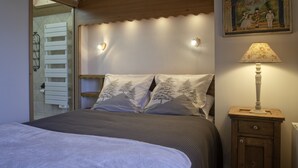 Ein ordentlich bezogenes Bett mit weißer Decke und zwei Kissen mit Baummotiven, ein Nachttisch aus Holz mit einer Lampe und ein an der Wand befestigtes Regal mit einem Bild.