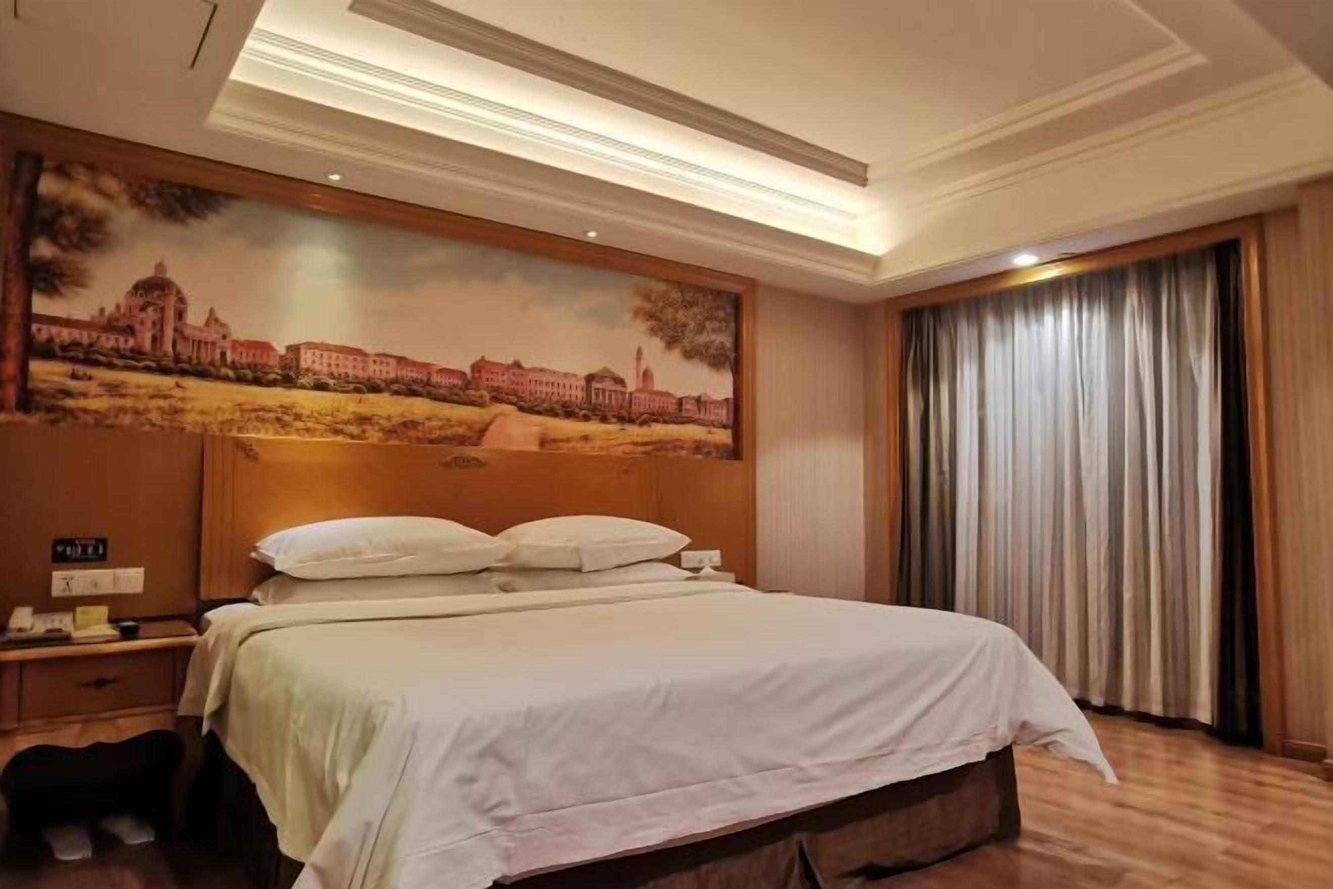 A hotel room with a large bed, a nightstand with a phone, a painting of a building on the wall, and a window with curtains.