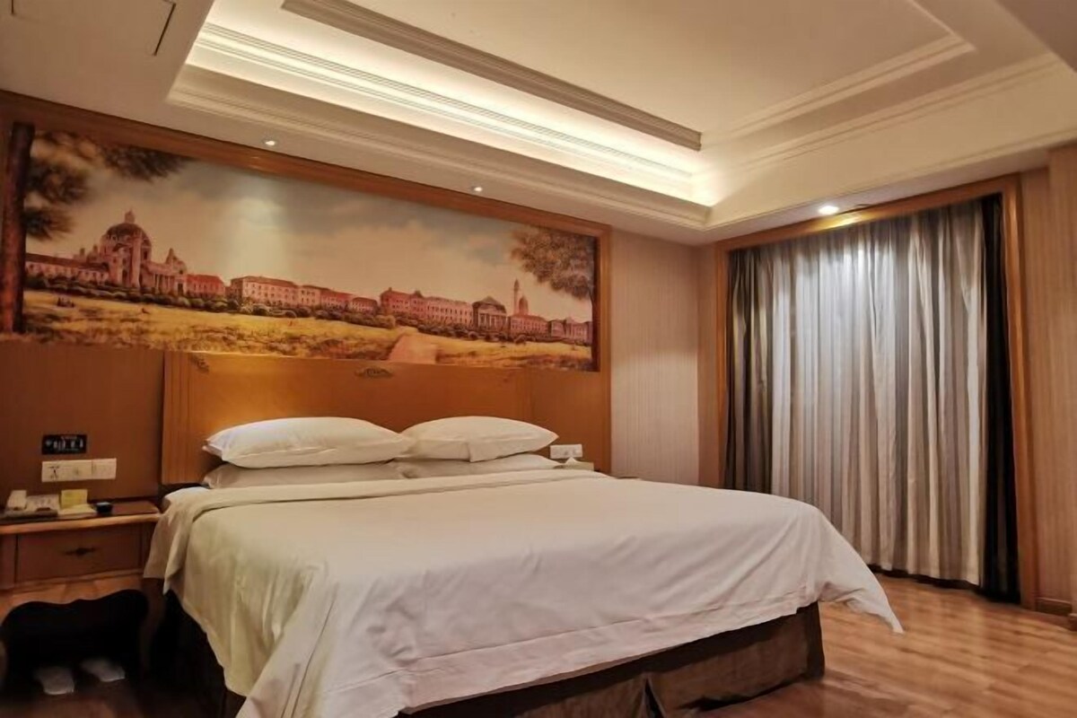 A hotel room with a large bed, a nightstand with a phone, a painting of a building on the wall, and a window with curtains.