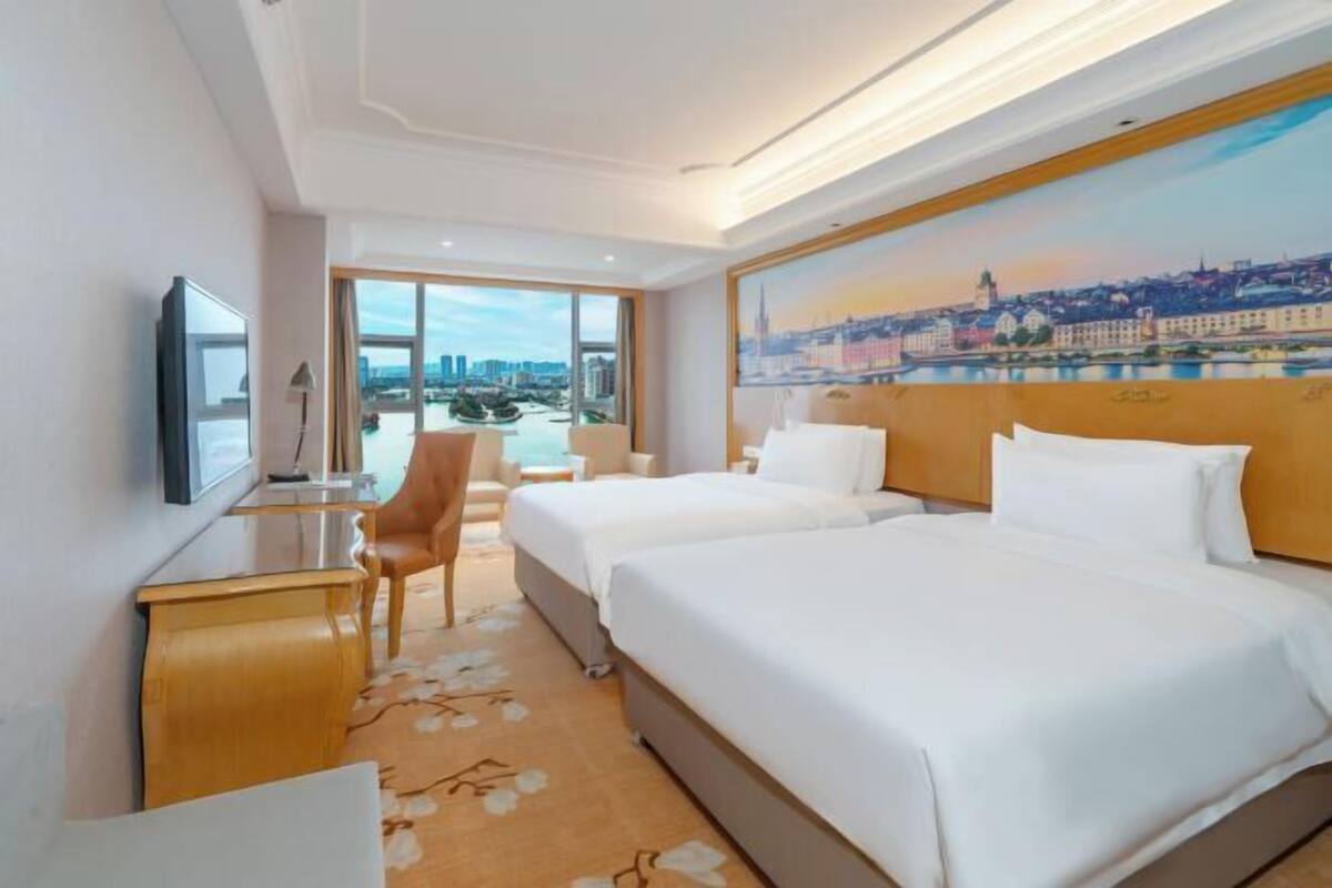 Habitación de hotel con dos camas, un escritorio, una silla y un amplio ventanal con vistas a la ciudad.