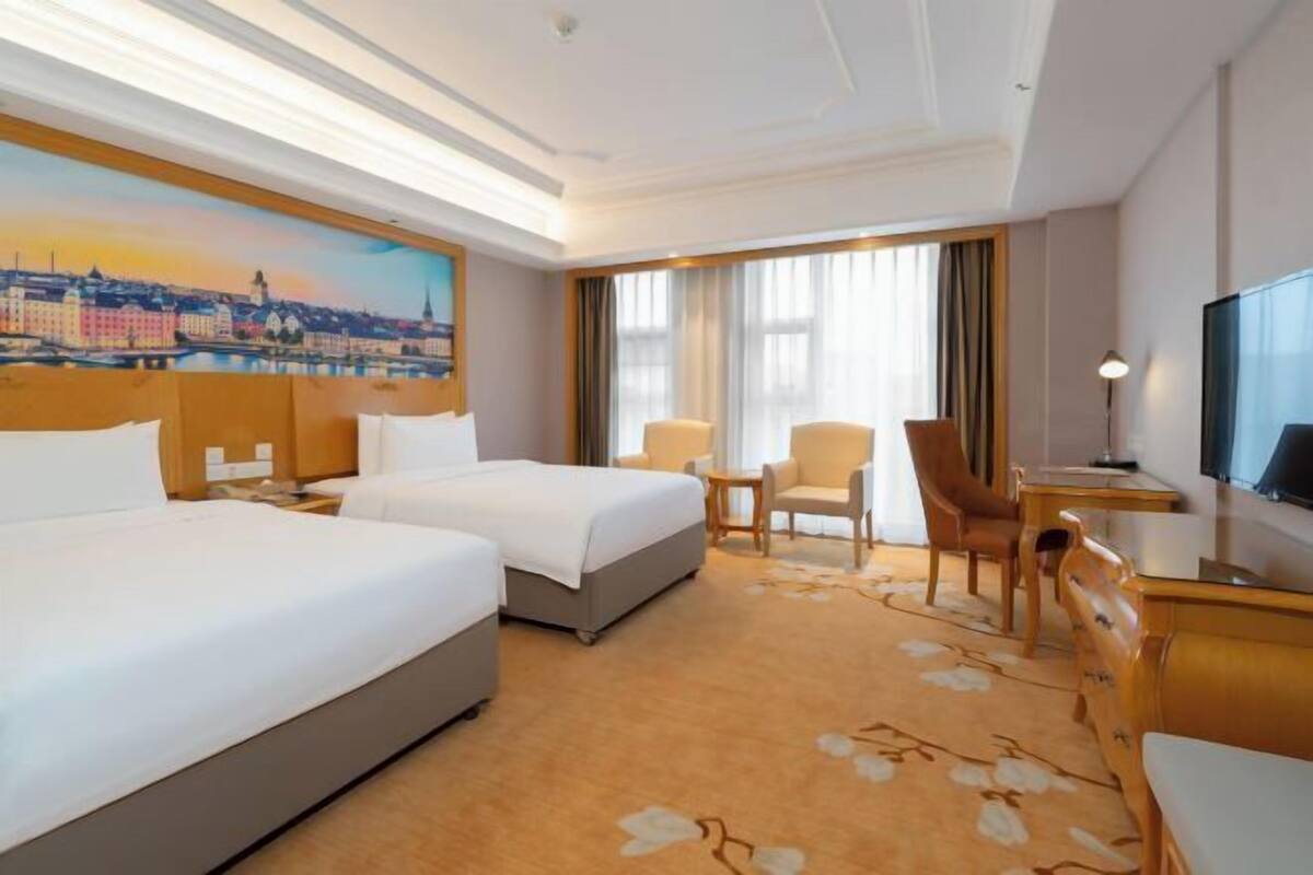 Habitación de hotel con dos camas, un escritorio, una silla, un televisor y un cuadro de un paisaje urbano.