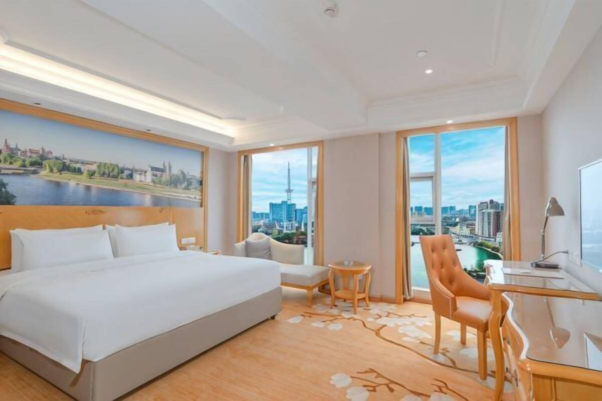 Habitación de hotel con una cama grande, un escritorio, una silla, una mesita y vistas a un paisaje urbano a través de amplios ventanales.