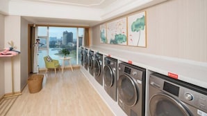 A laundry room with multiple washing machines, a small seating area with a chair, and a view of the city through large windows. - Vienna International Hotel (Tianmen Wanda Plaza Donghu Branch) (Tianmen)
