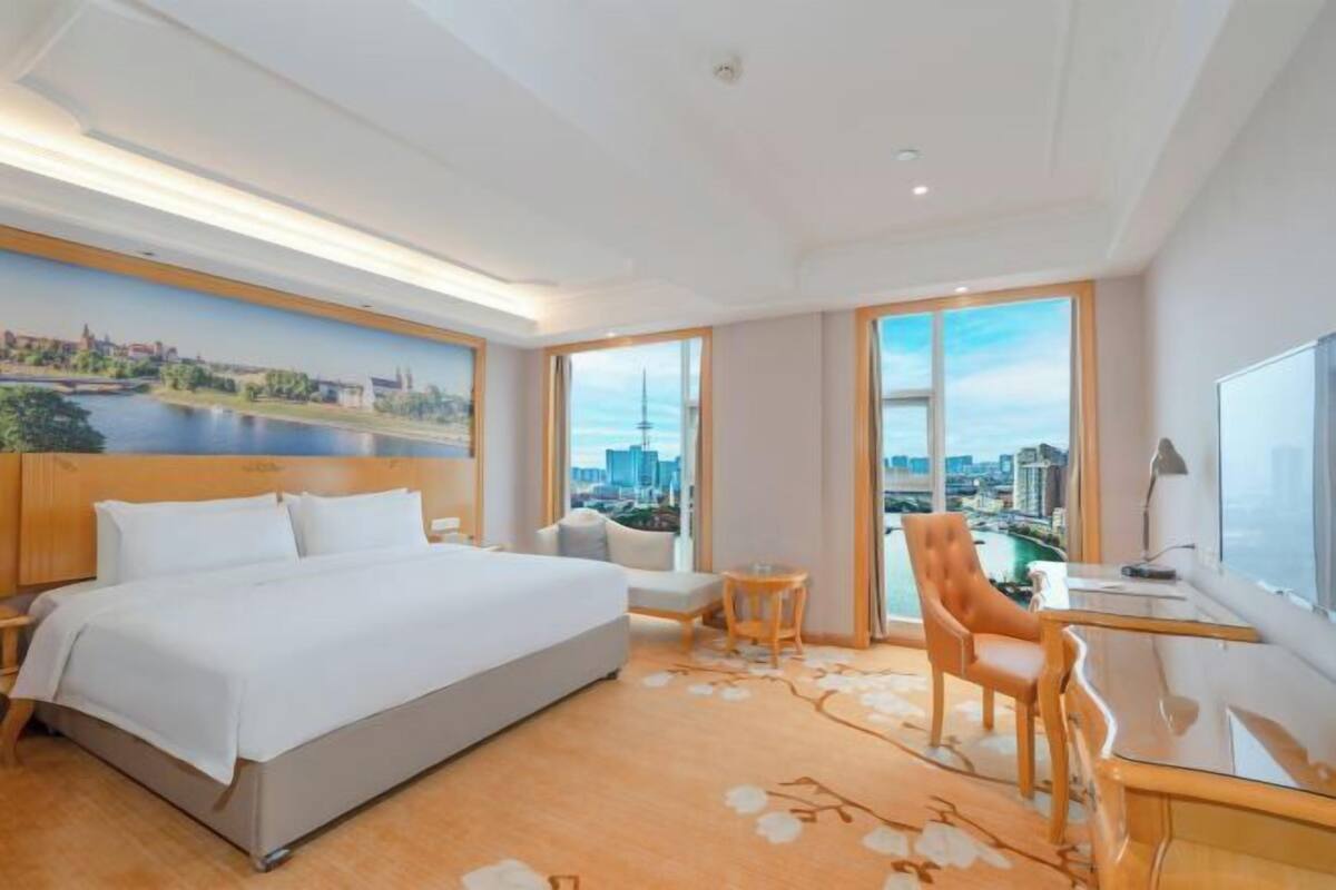 Habitación de hotel con una cama grande, un escritorio, una silla, una mesita y vistas a un paisaje urbano a través de grandes ventanales.