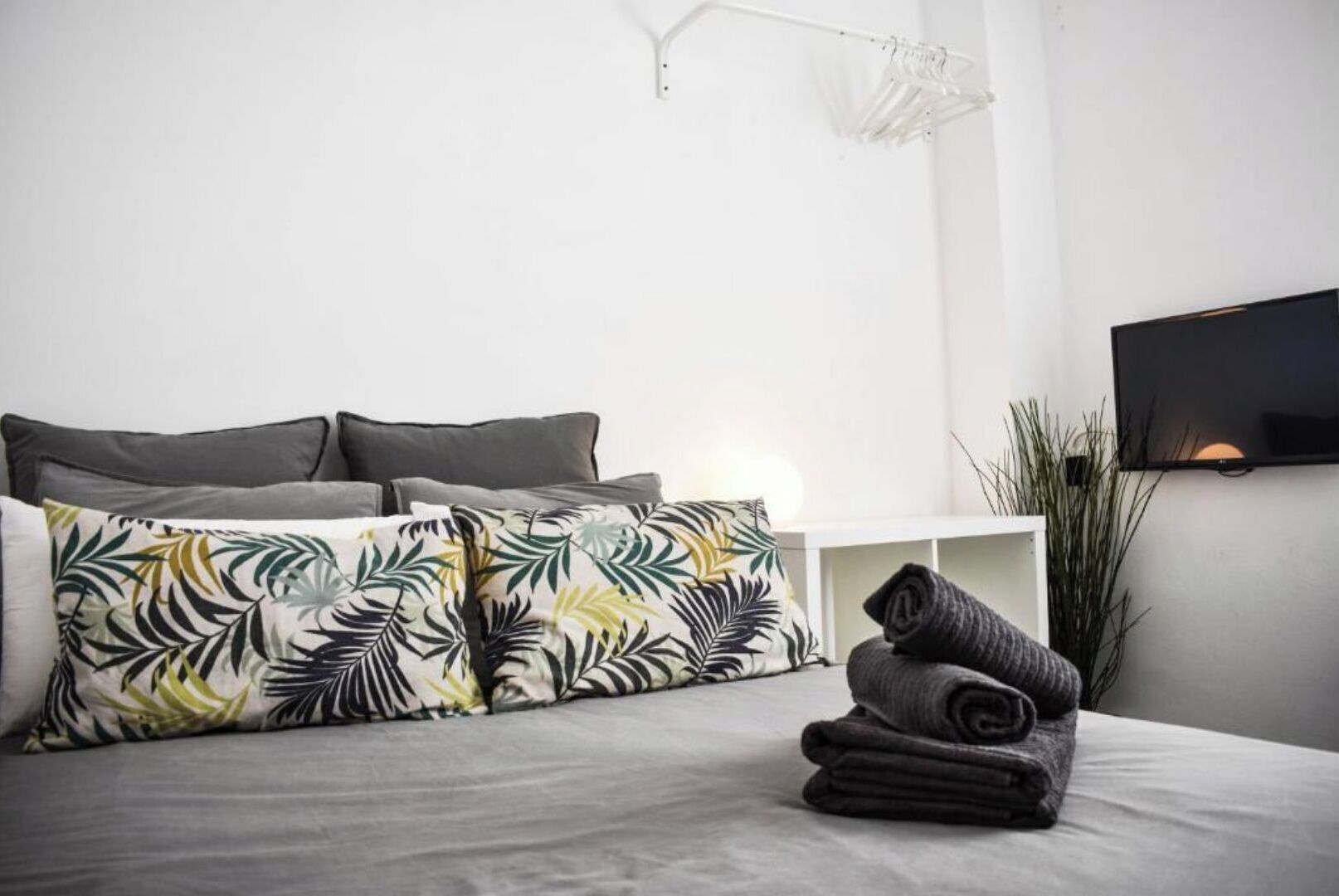Un dormitori modern amb un llit gris, almoinades amb dibuixos de fulles tropicals, una taula de nit blanca i una televisió penjada a la paret.