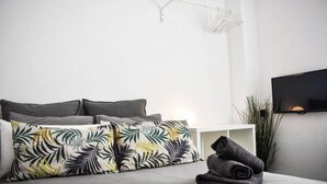 Quarto moderno com cama cinza, almofadas estampadas com folhagem tropical, mesa de cabeceira branca e TV fixada na parede.