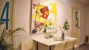 Une pièce avec une table et des chaises blanches, un tableau abstrait aux couleurs vives accroché au mur, et un panneau indiquant le mot « MAISON » posé sur la table.