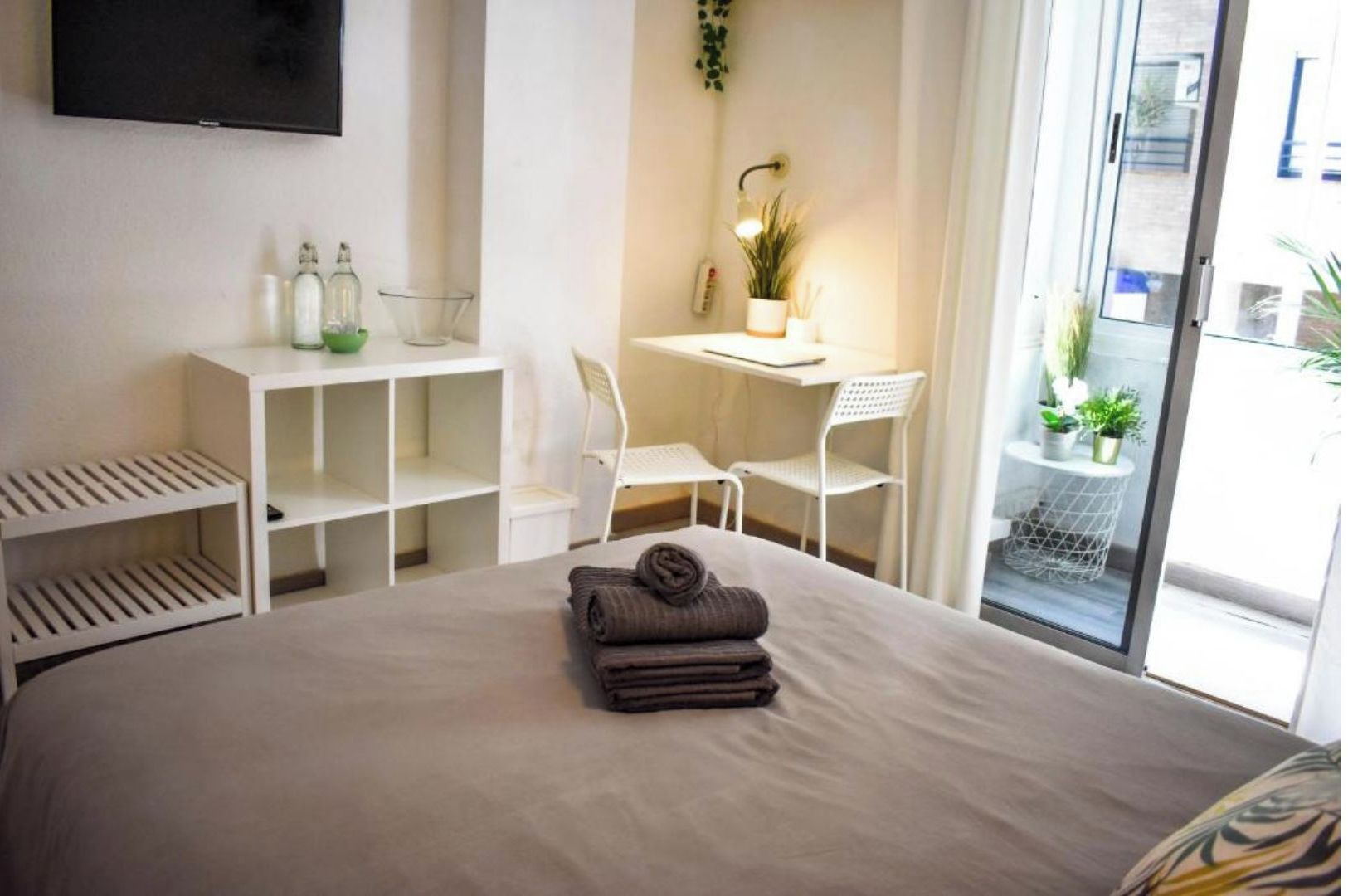 Une chambre d’hôtel avec un lit, un bureau, deux chaises, une télévision et un balcon agrémenté de plantes.