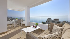 A balcony with a white table, chairs, and a sofa. There is a view of the sea and buildings in the background.