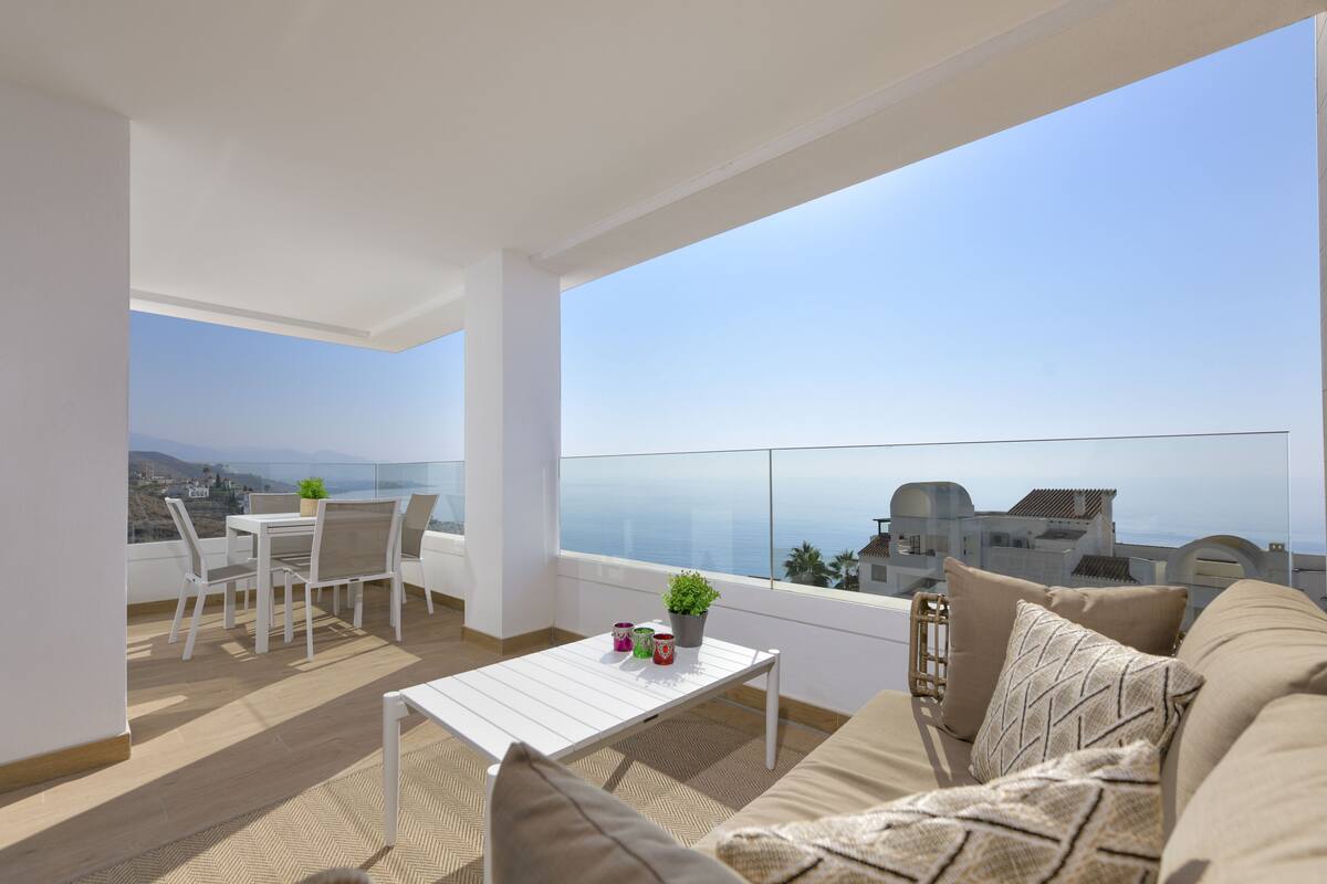 A balcony with a white table, chairs, and a sofa. There is a view of the sea and buildings in the background.