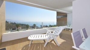 A balcony with a view of the sea, two white lounge chairs, and a glass table.