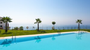 A swimming pool with clear blue water, surrounded by a fence and palm trees, with a view of the sea in the background.