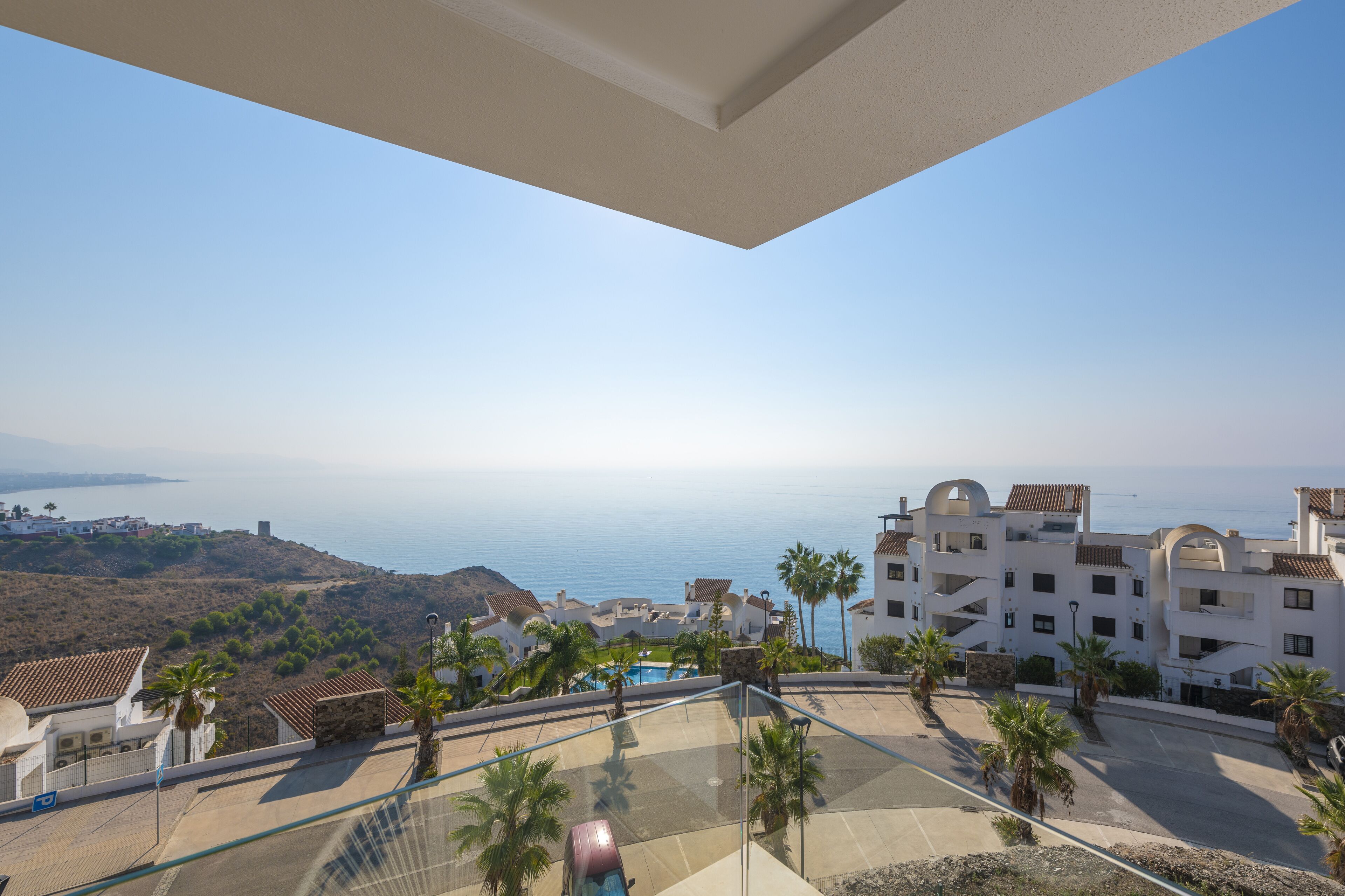A coastal view from a balcony overlooking a pool area with palm trees, a clear sky, and buildings with Mediterranean architecture.
