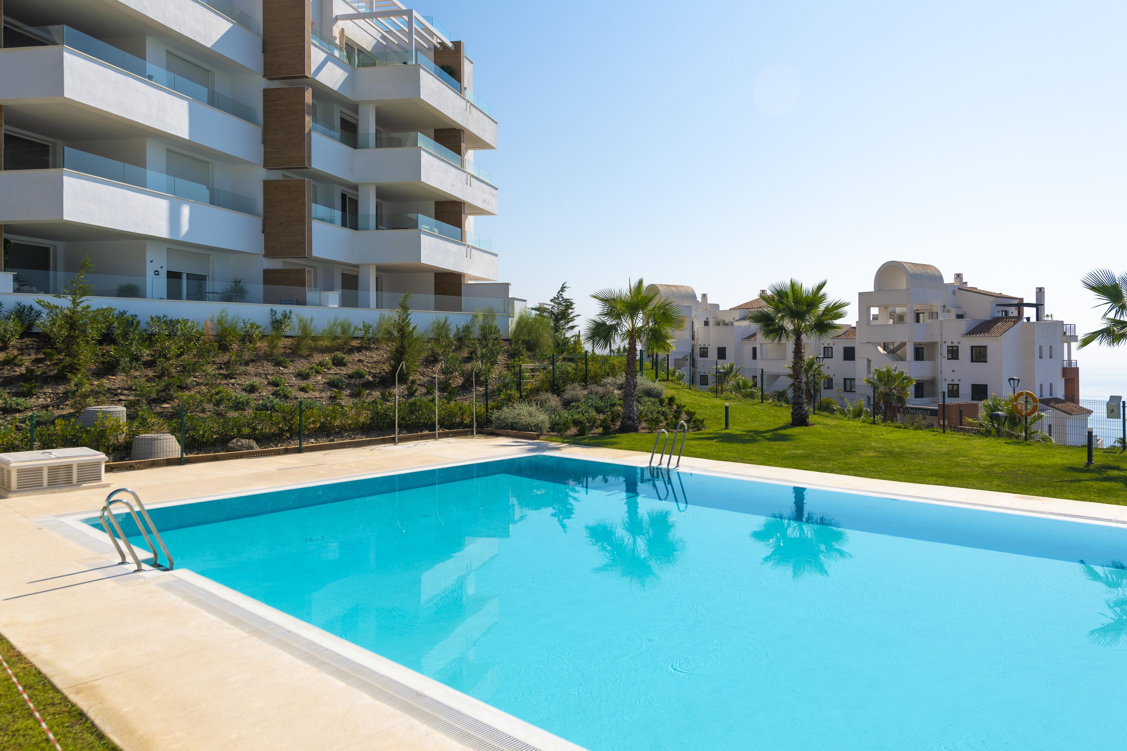 A swimming pool with a clear blue water, surrounded by a concrete deck. There are multiple balconies on the adjacent buildings, and palm trees are visible in the background.