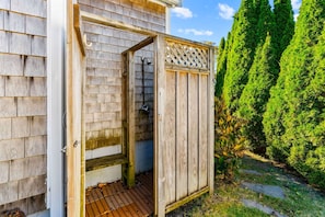 A wooden outdoor shower enclosure with a metal grate floor, adjacent to a house with siding and a wooden fence.