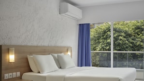 Una habitación de hotel moderna con una cama grande, una mesita de noche, aire acondicionado de pared y una ventana con vistas a árboles.