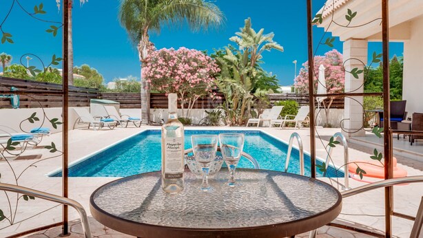 A poolside area with a glass table, lounge chairs, and a bottle of alcohol. - Villa Hanna  3 Bedrooms (Protaras)