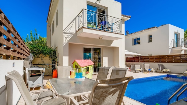 A poolside area with a table and chairs, a small playhouse, and a building with a balcony.
