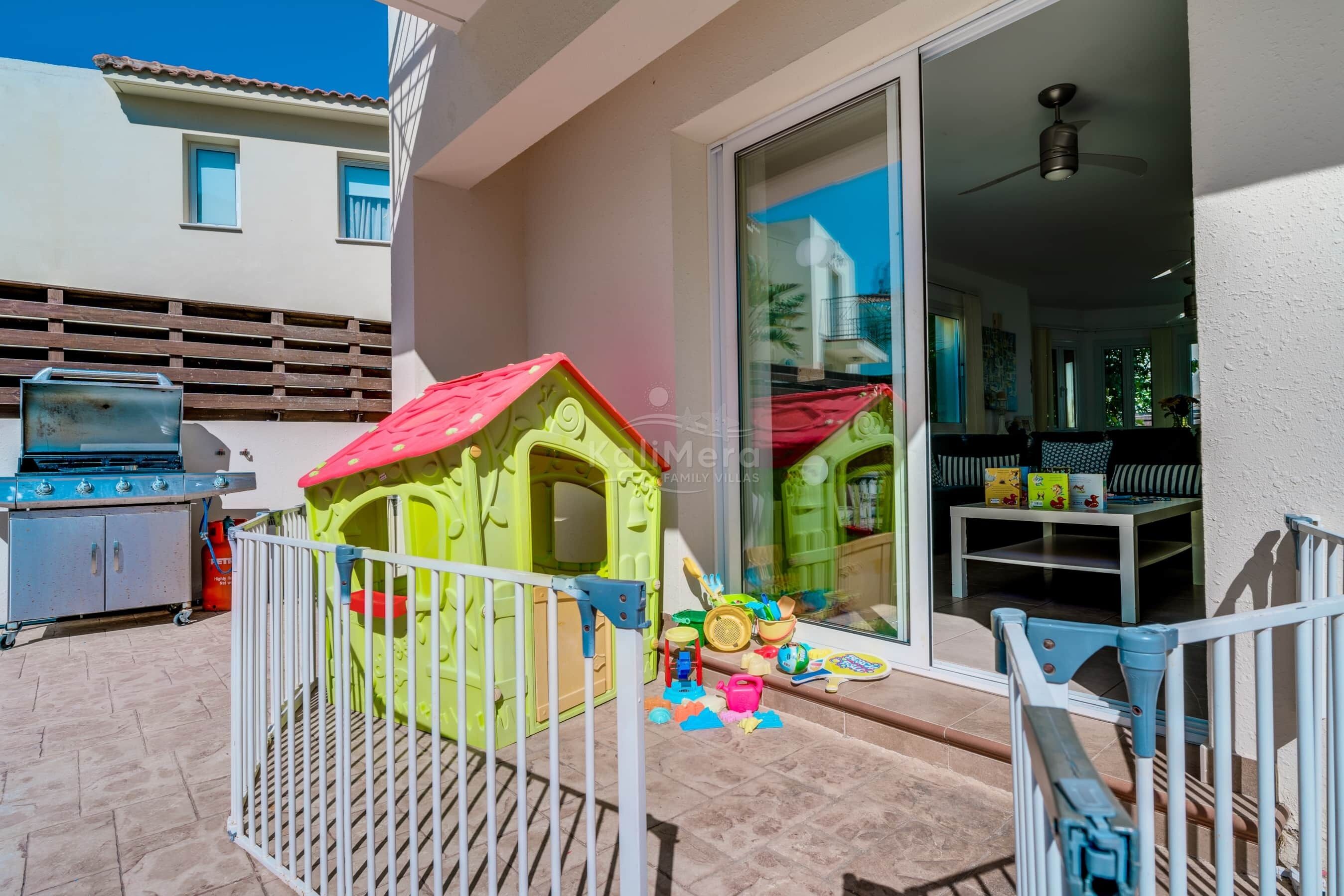 A patio with a playhouse, a barbecue grill, and a sliding glass door leading to an interior with a ceiling fan and seating area.