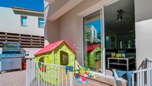 A patio with a playhouse, a barbecue grill, and a sliding glass door leading to an interior with a ceiling fan and seating area.