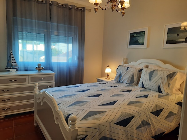 A bedroom with a bed, a nightstand, a dresser, a window with curtains, and a chandelier.