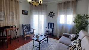 Ein Wohnzimmer mit Sofa, Sesseln, einem Couchtisch, einer Deckenleuchte und einem Essbereich mit Tisch und Stühlen.