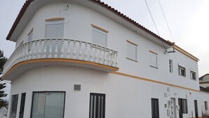 Ein zweistöckiges Gebäude mit weißer Fassade und orangenem Zierstreifen, das einen Balkon mit Brüstung, mehrere Fenster mit Rollläden und einen gefliesten Eingangsbereich aufweist.