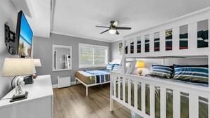 A bedroom with a bunk bed, a desk with a computer, a ceiling fan, and a window with blinds.